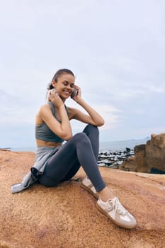 A joyful young woman in athletic wear listens to music with headphones, showcasing a healthy lifestyle and positive emotions against a scenic outdoor backdrop, illustrating fitness and relaxation