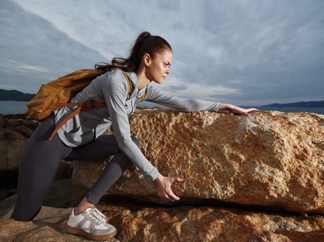 Determined athletic woman, dressed in stylish sportswear with a backpack, climbing a rock at the beach This image embodies strength, adventure, and outdoor fitness