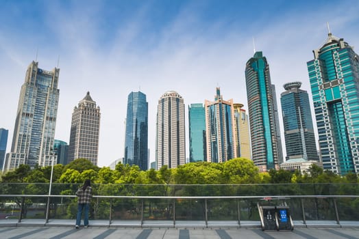 Shanghai, China - 1 April 2025: Modern Pudong business district with striking skyscrapers of diverse architecture against blue sky. A person admires the view from glass railings above lush urban park