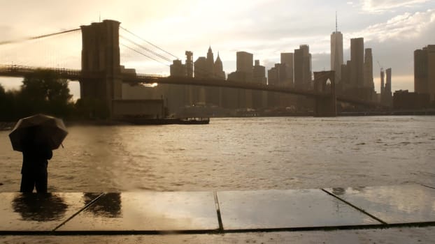 New York City, United States - 11 Sept 2023: Brooklyn Bridge rainy foggy day. Manhattan downtown skyline cityscape, World Trade Center. Man with umbrella, Dumbo Pebble Beach. Autumn river, fall in USA