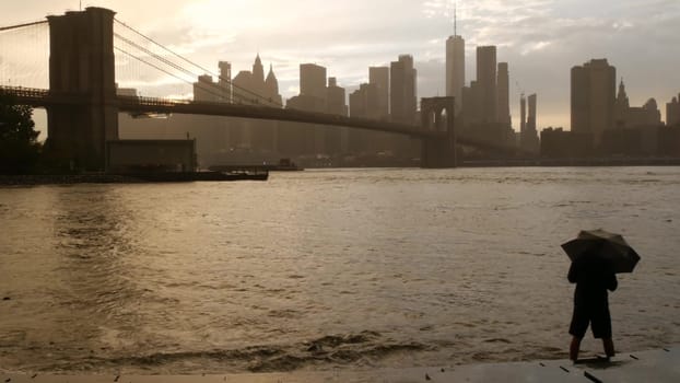 New York City, United States - 11 Sept 2023: Brooklyn Bridge rainy foggy day. Manhattan downtown skyline cityscape, World Trade Center. Man with umbrella, Dumbo Pebble Beach. Autumn river, fall in USA