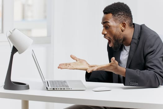 A businessman in a suit sits at a desk with a laptop, mouth open in shock, hands resting on the desk.