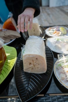 Hand of a seller and a piece of cheese in a transparent package at a farmers' market. Natural cheese at a city fair. A large piece of cheese on a black oval plate.
