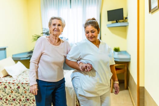 Nurse supporting elderly woman while walking in a nursing home room, providing care and assistance