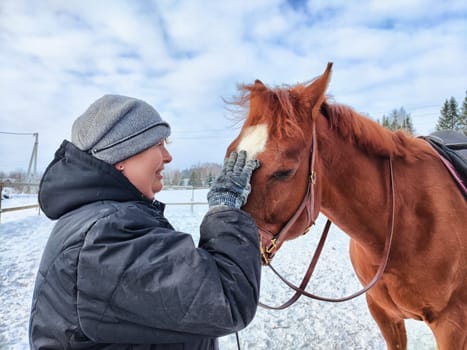 In a snowy setting, a woman gently pets a chestnut horse, enjoying a warm moment of connection during winter.