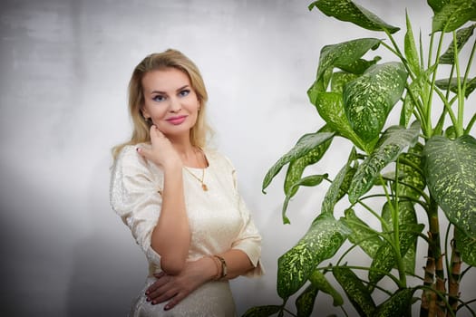 A middle-aged woman styled as a blogger poses elegantly indoors next to a lush plant, showcasing her lifestyle.