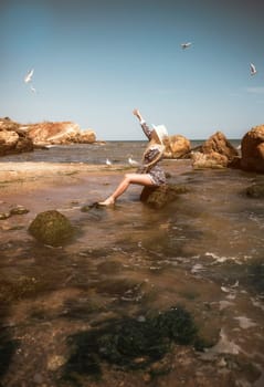 young blonde in a short dress and hat sits on a rock in the sea on the beach. her hair flutters in the wind, she enjoys the sun, seagulls fly around her general plan