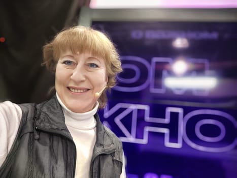 A middle-aged woman smiles for a selfie while attending a lively tourism event focused on travel and lifestyle blogging.