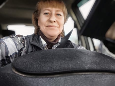 A middle-aged woman takes a selfie while driving, sharing her travel experience on her lifestyle blog during a scenic road trip.