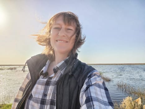 Blonde woman takes a cheerful selfie during her adventure by the water, soaking in the sun and enjoying the peaceful surroundings.