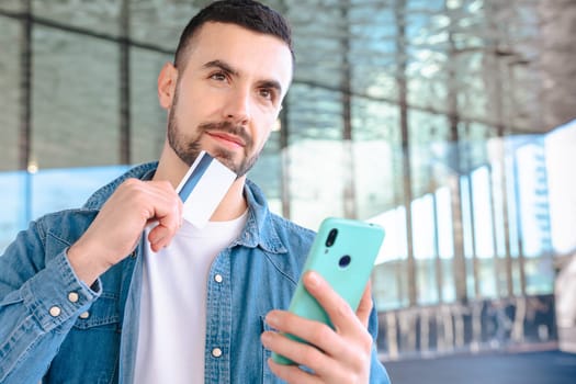 Happy young man using his smartphone while holding a credit card. He smiles with excitement, standing outside a glass building, enjoying an online shopping or payment experience.