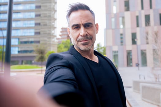 Caucasian man with confident expression takes a selfie in a modern city environment. Wearing black outfit, he stands among tall buildings. Urban lifestyle and personal branding concept.