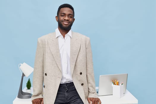 A cheerful man in a beige blazer and white shirt standing at his desk with a laptop, desk lamp, and writing utensils, captured in a bright modern office with a light blue background, conveying professionalism and approachability. Concept: modern workplace wellness