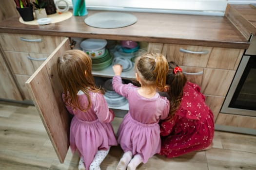 In a warm kitchen filled with natural light, three young girls in lovely dresses enthusiastically arrange vibrant plates from a cabinet.