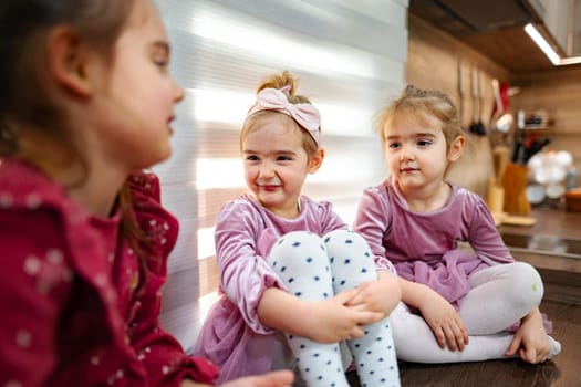 In a bright kitchen filled with warmth, three young girls engage in cheerful conversation. Dressed in soft, matching outfits, they share smiles and laughter, creating a joyful atmosphere.