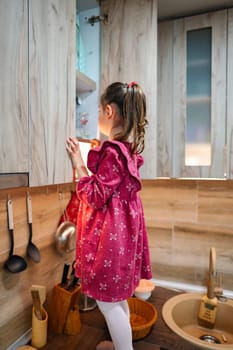 In a warm kitchen setting, a young girl in a pink dress reaches for something on a shelf, showcasing her curiosity and adventurous spirit as she discovers her surroundings.