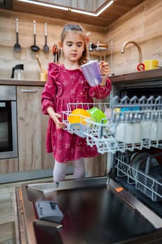 A young child in a bright pink dress carefully loads colorful dishes into a dishwasher in a modern kitchen. Soft light illuminates the wooden cabinetry, highlighting her focused expression.