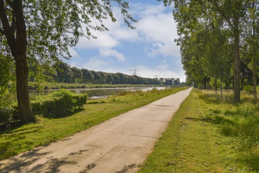 A tranquil pathway lined with trees, leading towards a river under a bright, partly cloudy sky. Ideal for walking and enjoying nature's beauty.