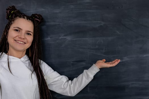 A cheerful young woman happily displays a welcoming gesture beside an empty and blank chalkboard, inviting ideas