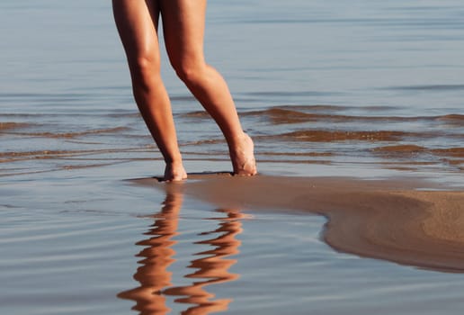 A nude woman on the seashore. A persons legs walking on the shoreline with a reflection on the wet sand, near the water.