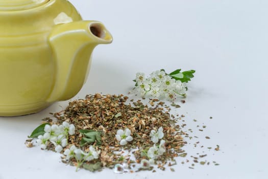 A yellow teapot sits next to a blend of dried herbs and crushed flowers on a light surface. Fresh white blossoms and green leaves are arranged nearby, highlighting the tea-making process.