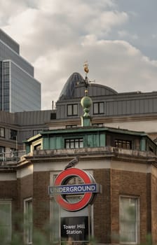 London, UK - Apr 27, 2025 - View of A pigeon perched on Underground sign at Tower hill station with Trinity House, Skyscrapers and sky in the background. use it as your Wallpaper, Poster and Space for text, Selective focus.