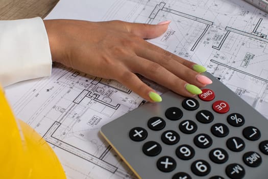A closeup of a hand pressing calculator buttons, surrounded by various architectural blueprints and plans