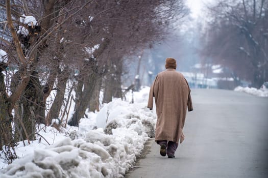 Srinagar, Kashmir - 4th Jan 2025: Man in traditional Kashmiri Phiran walking along a snow covered road with trees on both sides showing the harsh winters in the region