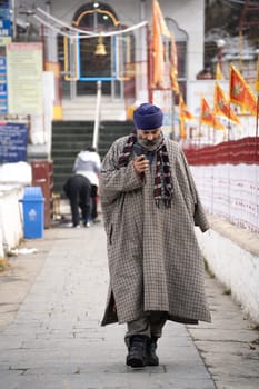 Srinagar, Kashmir - 4th Jan 2025: Sikh man wearning the traditional phiran overcoat cloak of kashmir walking out of a temple while talking on phone