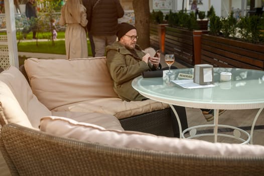 Odessa, Ukraine - April 21, 2025: A bearded man in glasses with a phone in his hands. He is relaxing in a cafe, outdoors, at a table with a glass of white wine. He is wearing a hat and a raincoat.