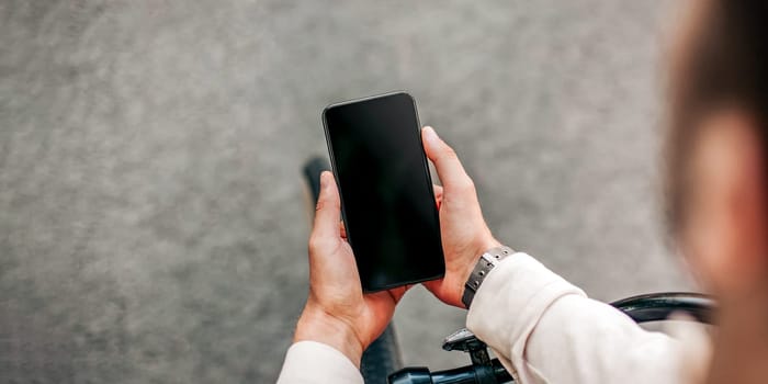 Social networks, work online, good app and modern lifestyle. Young man freelancer or blogger sits on bicycle and looks in smartphone with blank screen in hands, outdoor, cropped, free space