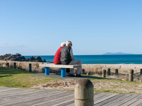 Mount Maunganui New Zealand - November 4 2012; Elderly couple with grey hair sit on bench looking at ocean view on sunny day.