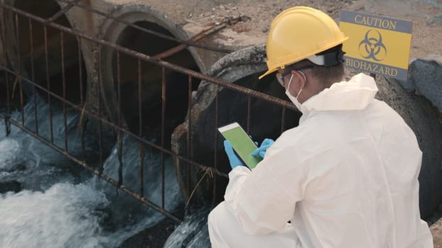Ecologist sampling water of toxic chemicals from river touching screen on digital tablet, Biologist wear protective suit and mask collects sample of waste water from industrial, problem environment