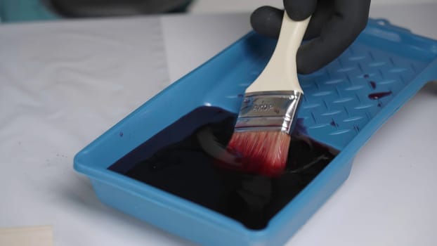 A paintbrush is being dipped into dark paint that resides in a blue tray, fully prepared for application. Creative