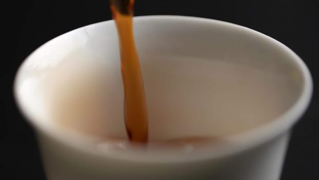 A closeup image of a steaming hot beverage being skillfully poured into a smooth white cup against a dark background. Media