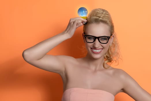 beautiful  blonde girl with pink dress and black glasses having an idea. She taking light bulb near head with funny expression