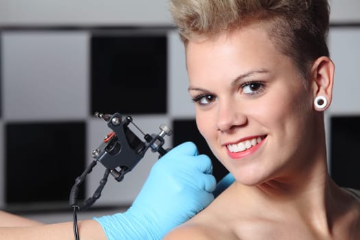 Beautiful woman looking at camera while somebody is making a tattoo on her shoulder
