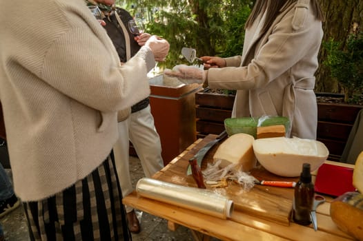 hands of cut unrecognizable women at a street fair, cheese trade. cheese tasting process. A vendor uses a slicer with a wooden handle to serve a customer a slice of cheese surrounded by cheese wheels