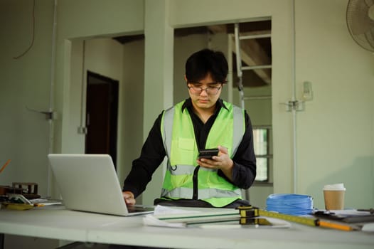 Engineer using smartphone at construction planning desk with tools and blueprints.