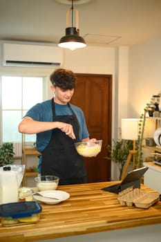 Young man mixing pancake batter in the kitchen while following a recipe on a digital tablet..