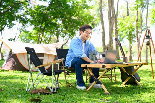 Young man using laptop at a table while camping in the forest.