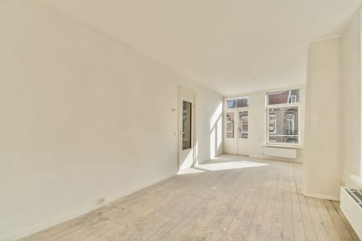A bright, spacious room featuring large windows that allow abundant natural light. The area has a minimalistic design with light-colored walls and wooden flooring.
