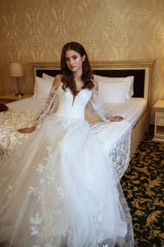 Young elegant brunette bride in vintage white dress posing in hotel before wedding. Long white dress with long sleeves, corset and open shoulders. Bride portrait, wedding makeup, fashionable bride