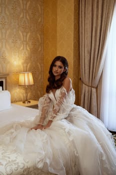 Young elegant brunette bride in vintage white dress posing in hotel before wedding. She is sitting on bed in hotel room. Long white dress with long sleeves, corset and open shoulders. wedding makeup