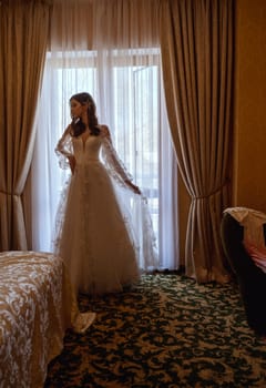 Bride in long traditional white dress standing near window, in hotel. Bride with long hair standing in front of window in hotel suite before wedding.
