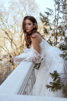 Young brunette bride in traditional long white dress posing on hotel balcony in spring. white dress with long sleeves, corset and open shoulders. hotel with classical architecture. fashionable bride