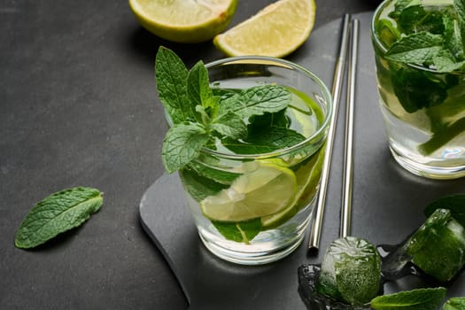 Glass of mojito cocktail with fresh mint leaves, lime slices, and ice cubes on a dark stone background. Refreshing summer drink with stainless steel straws