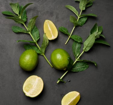 Whole and cut limes with fresh mint leaves arranged on a dark textured surface. Composition perfect for food, drink, or summer themes