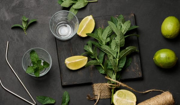Bunch of fresh green mint on wooden board and lime wedges, glasses and metal straws, top view. Cocktail ingredient