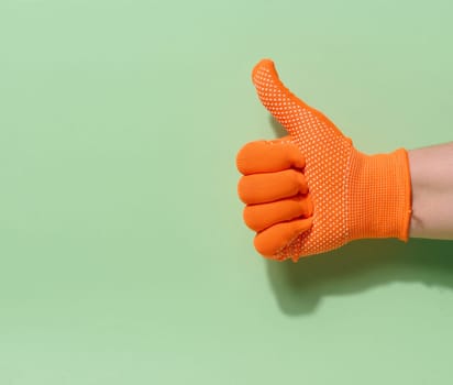 A hand wearing a bright orange work glove with small white knobs gives a thumbs up gesture against a green background with ample copy space
